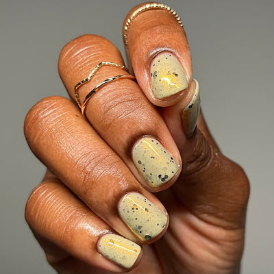 OG Devo-tea by Dam Nail Polish, warm pale sage-tan crelly with gold shimmer streaks and speckled black glitter, on a medium-deep skin tone hand