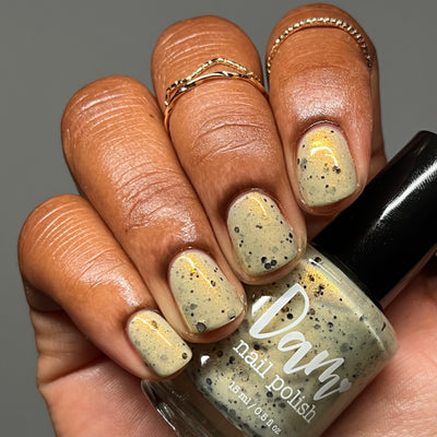OG Devo-tea by Dam Nail Polish, pale tan-cream crelly with gold shimmer flashes and assorted black glitter particles, bottle held in a medium-deep skin tone hand