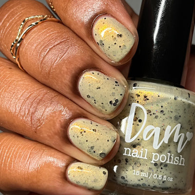 OG Devo-tea by Dam Nail Polish, pale cream-tan crelly with gold shimmer and scattered black glitters of varying sizes, bottle held in a deep skin tone hand