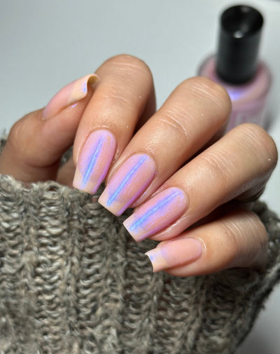 A Whimsical Dream by Dam Nail Polish, sheer milky pink base with linear blue-purple shimmer streaks over a lavender mid-tone, on a light-medium skin tone hand
