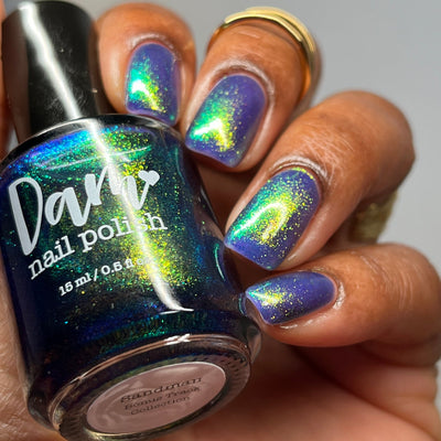 Sandman by Dam Nail Polish, blurple jelly with vivid gold-green-blue color-shifting shimmer, bottle held in a medium-deep skin tone hand