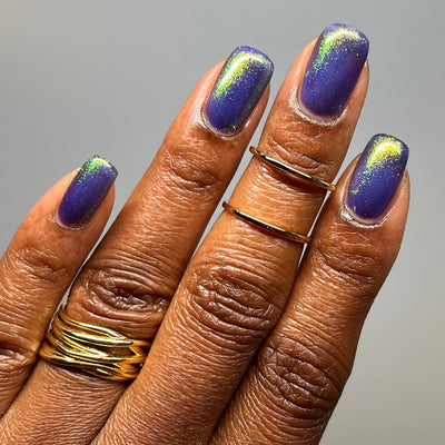 Sandman by Dam Nail Polish, cool blurple jelly with vivid yellow-green and teal color-shifting shimmer concentrated near cuticle and tip, on a deep skin tone hand