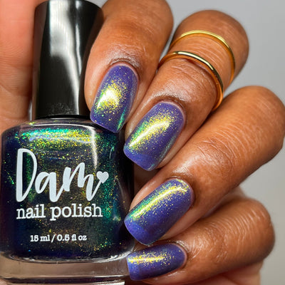 Sandman by Dam Nail Polish, cool blurple jelly showing vivid gold, green, and blue color-shifting flakies, bottle held in a medium-deep skin tone hand