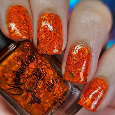 Sauron's Eye by Chamaeleon Nails, fiery orange jelly with copper-to-olive UCC flakies and deep amber-red iridescent flakes in bright daylight, on a light skin tone hand
