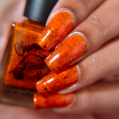 Sauron's Eye by Chamaeleon Nails, vivid red-orange jelly with copper-to-green UCC flakies in bright daylight, bottle held in a light-medium hand