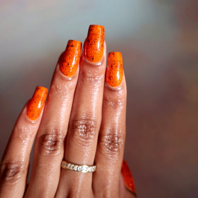 Sauron's Eye by Chamaeleon Nails, fiery orange jelly with scattered dark copper and iridescent UCC flakies showing deep flakie texture, on a medium skin tone hand