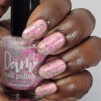 Shimmer Storm by Dam Nail Polish, sheer warm white crelly base with neon pink shimmer and scattered pink and dark brown-black glitter, bottle held in a rich skin tone hand