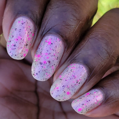 Shimmer Storm by Dam Nail Polish, sheer white base with multitonal neon pink and dark mauve glitter flakes intensified in bright daylight, on a rich skin tone hand