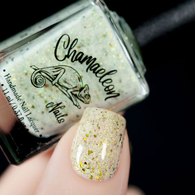The Forgotten Lagoon (UV Polish) by Chamaeleon Nails, cream-white flakie UV in resting state with gold-green and orange shifting multichrome and UCC flakes under studio light, bottle held in a fair hand