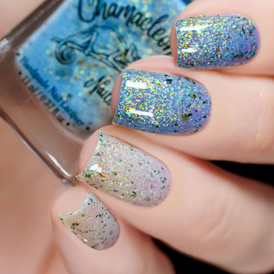 The Forgotten Lagoon (UV Polish) by Chamaeleon Nails, flakie UV showing dramatic UV shift from sheer white-lavender base to periwinkle blue-lavender in bright daylight, with dense green-gold-bronze iridescent flakes, on a light skin tone hand