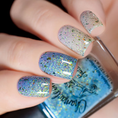The Forgotten Lagoon (UV Polish) by Chamaeleon Nails, flakie UV showing white-to-periwinkle blue base gradient across nails, with large green-to-gold multichrome and orange iridescent flakes, on a light skin tone hand
