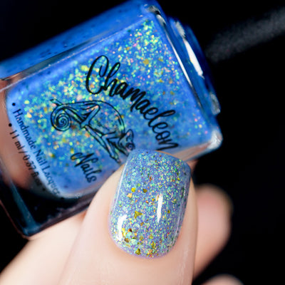 The Forgotten Lagoon (UV Polish) by Chamaeleon Nails, periwinkle blue flakie UV in UV-activated state with green-to-gold iridescent flakes and large bronze ultrachrome shards, bottle held in a light hand