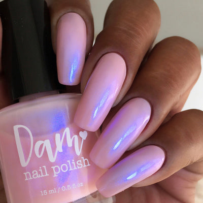 A Whimsical Dream by Dam Nail Polish, sheer pink shimmer with vivid blurple iridescent shift in bright daylight, bottle held in a medium-deep skin tone hand