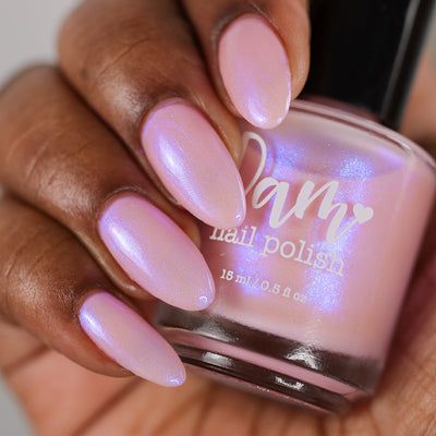 A Whimsical Dream by Dam Nail Polish, sheer baby pink base with vivid blue-purple shimmer pooling under studio light, bottle held in a deep skin tone hand