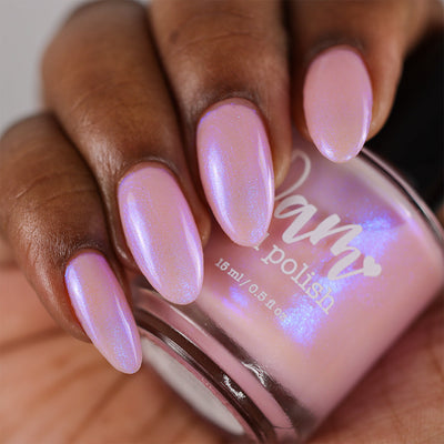 A Whimsical Dream by Dam Nail Polish, sheer soft pink base with blurple shimmer shifting to violet-blue under studio light, bottle held in a deep skin tone hand
