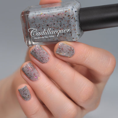 Cadillacquer Daydreaming grey red chameleon iridescent crelly nail polish, bottle held with four-nail swatch, light skin, studio lighting