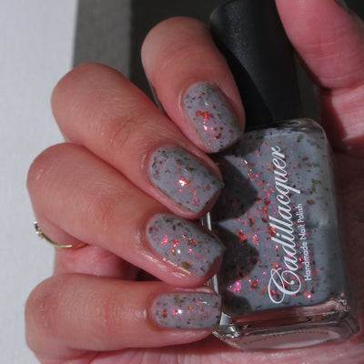 Cadillacquer Daydreaming grey red chameleon iridescent crelly nail polish, bottle held with four-nail swatch, light skin, indoor warm lighting