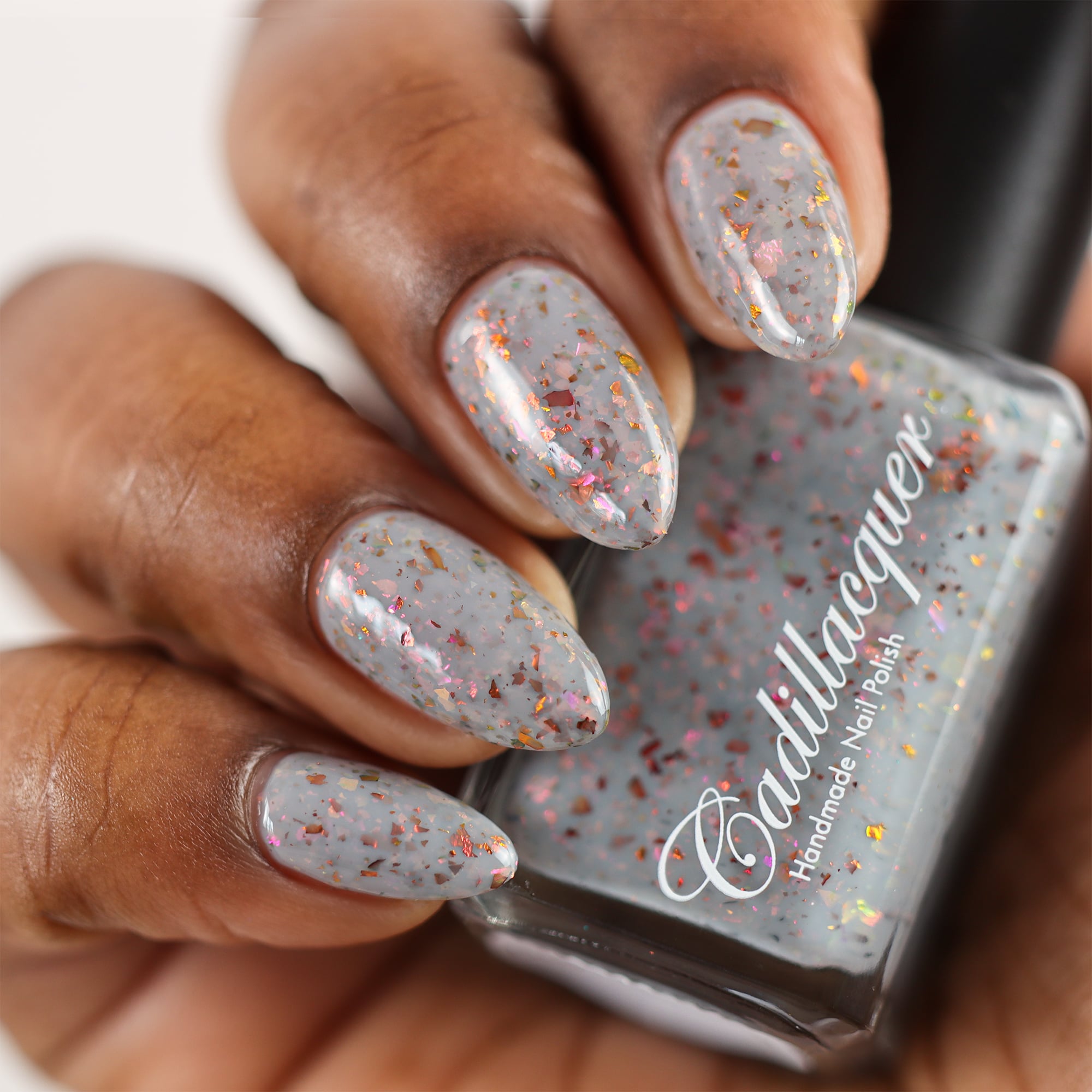 Cadillacquer Daydreaming grey red chameleon iridescent crelly nail polish, bottle held with four-nail swatch, deep skin, studio lighting