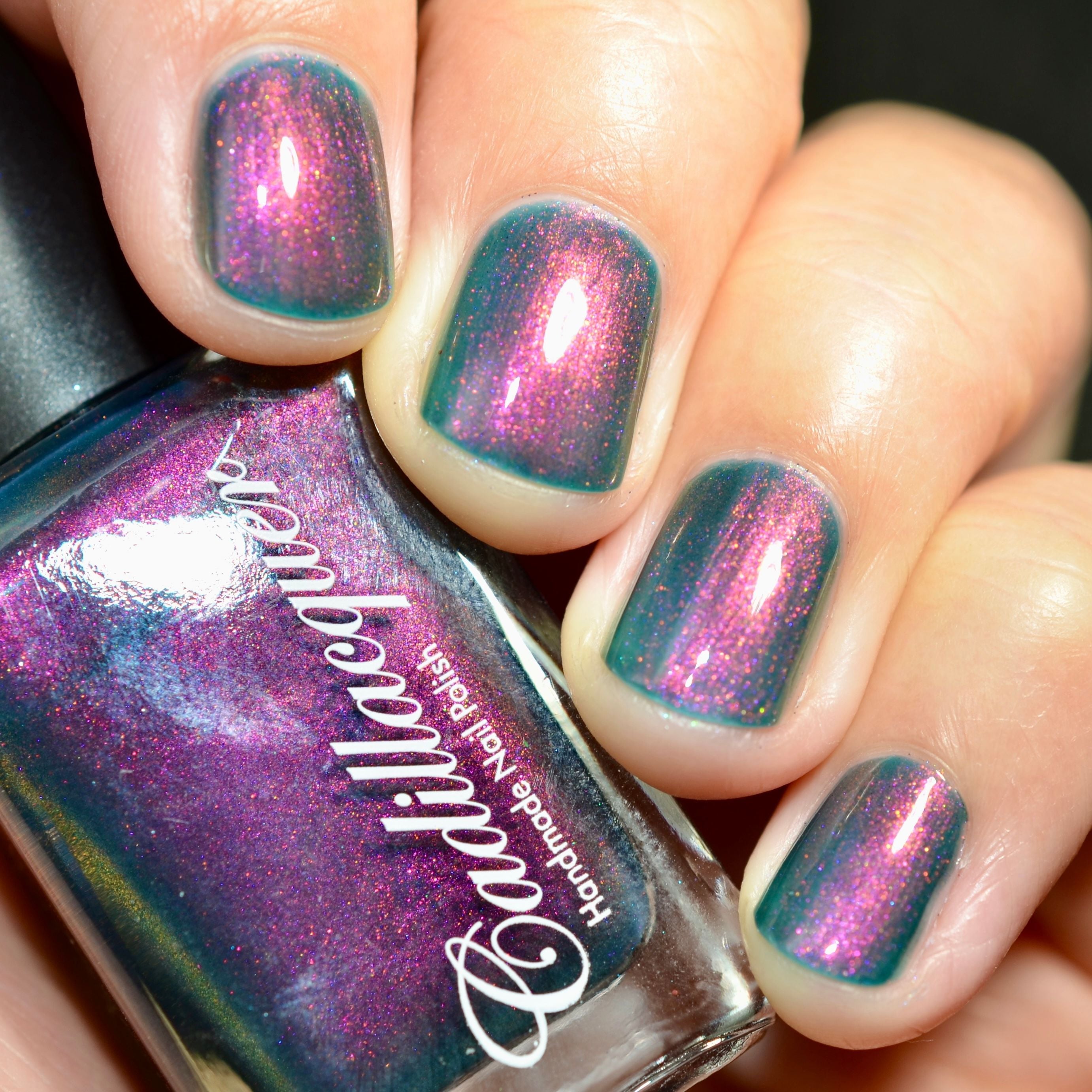 Multichrome shimmer Cadillacquer nail polish in Fear What You Will Become, bottle held in hand beside four swatched squoval nails shifting from murky teal base through magenta to orange shimmer, light skin tone, studio lighting, swatched by @caitlinswatches