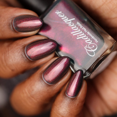 Multichrome shimmer Cadillacquer nail polish in Fear What You Will Become, shown on eight almond-shaped nails on a rich skin tone hand holding the polish bottle, the polish reads deep near-black burgundy in shadow shifting to vivid magenta and warm rose-pink in the light, with micro shimmer sparkling copper-orange to magenta at varying angles, studio lighting, swatched by @doseoflolade