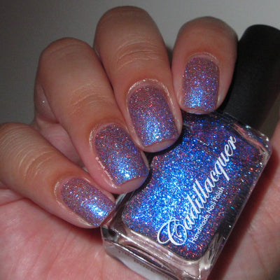 Shimmer, flake, and glitter Cadillacquer nail polish in I'll Be Right Back, shown in bottle held in hand with five swatched squoval nails, violet-purple sheer base densely packed with blue micro flakes and blue shimmer producing vivid cerulean flash, scattered red and charcoal reflective glitter sparks, light-medium skin tone, direct flash lighting, swatched by @_always_polished_