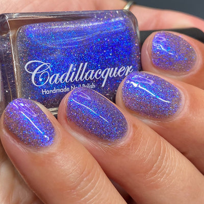 Holographic shimmer and micro-flake Cadillacquer nail polish in I'll Be Right Back, shown as bottle-in-hand with four swatched oval nails, polish is a dense blue-violet packed with blue micro flakes, royal blue shimmer flash, and scattered red and charcoal holographic glitter, light-medium skin tone, studio lighting, swatched by @heeymercedes