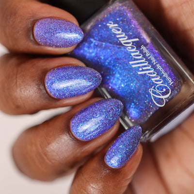 Shimmer, flakies, and reflective glitter Cadillacquer nail polish in I'll Be Right Back, shown on eight almond-shaped nails held around the polish bottle, shifting from blue-violet to bright cobalt blue with electric blue micro flakes and scattered charcoal and red reflective glitter, deep skin tone, studio lighting, swatched by @doseoflolade