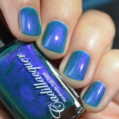 Multichrome shimmer Cadillacquer nail polish in It's All For You, shown on four short natural nails with a teal green base that shifts to violet-purple at angles, alongside the bottle which reads deep purple on its face with a teal base colour at the rim, on light skin tone under studio lighting, swatched by @caitlinswatches