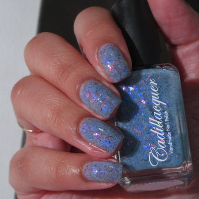 Cadillacquer Just Breathe pastel blue iridescent crelly nail polish, bottle held with five-nail swatch, light skin, indoor warm lighting