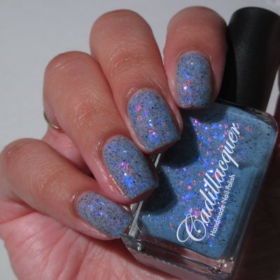 Cadillacquer Just Breathe pastel blue iridescent crelly nail polish, bottle held with four-nail swatch, light-medium skin, indoor warm lighting