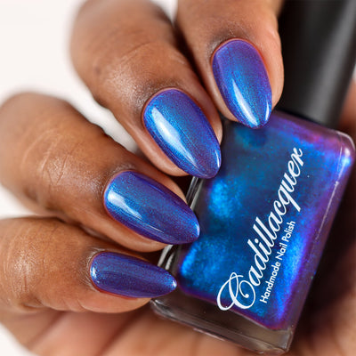 Multichrome shimmer Cadillacquer nail polish in Keep Doubting, shown on five almond-shaped nails on rich skin tone alongside the bottle held in hand, shifting from deep mulberry-violet base to vivid electric blue and teal-green shimmer flash depending on angle, studio lighting, swatched by @doseoflolade