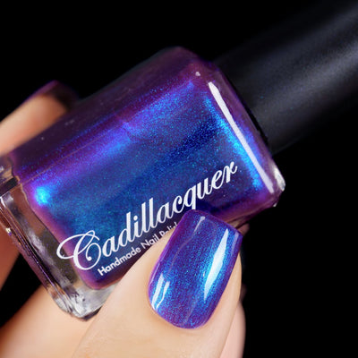 Multichrome shimmer Cadillacquer nail polish in Keep Doubting, shown as a bottle held in hand beside one swatched square nail, the bottle face shifts dramatically from deep violet through electric blue to teal-green shimmer while the nail reads medium purple-violet with blue shimmer flash, light skin tone, studio lighting against black background, swatched by @yyulia_m