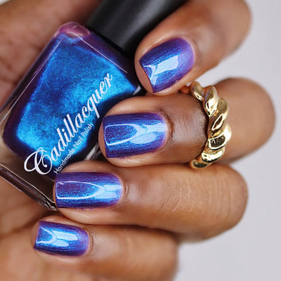 Shimmer multichrome Cadillacquer nail polish in Keep Doubting, shown in bottle held in hand with four swatched square nails shifting from deep blue-violet to electric cobalt blue, worn on deep skin tone with a gold twisted ring, studio lighting, swatched by @lacquer_is_life