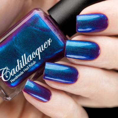 Multichrome shimmer Cadillacquer nail polish in Keep Doubting, shown as a bottle held against four swatched squoval nails on light skin, the polish shifts from deep mulberry-purple through electric cobalt blue to teal depending on viewing angle, with densely packed reflective blue shimmer particles, studio lighting, swatched by @rafinails