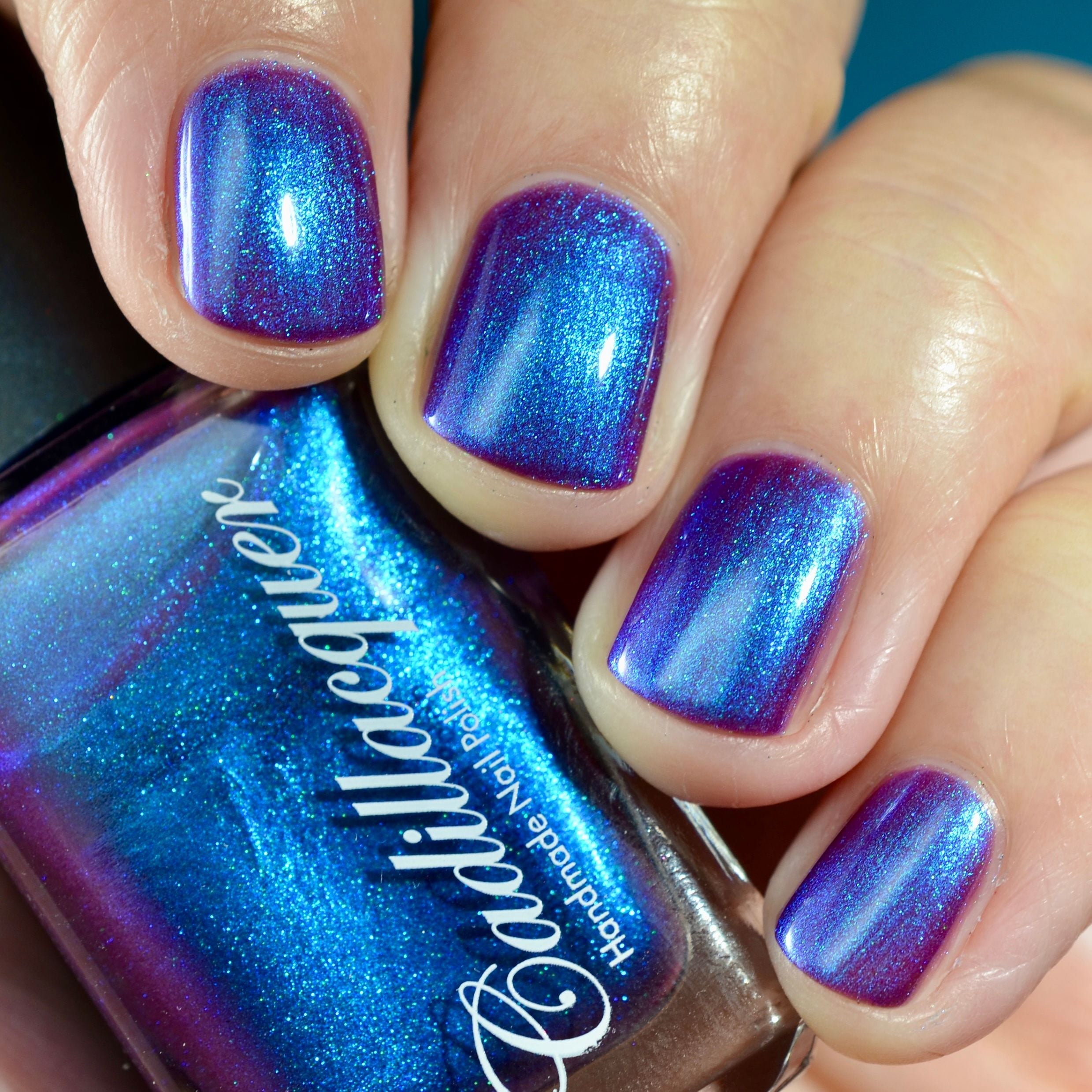 Multichrome shimmer Cadillacquer nail polish in Keep Doubting, shown swatched on four short natural nails held around a Cadillacquer bottle, nails shift from deep violet-purple at face-on angles to electric blue and mulberry-magenta at oblique angles with vivid blue shimmer throughout, light skin tone, studio lighting, swatched by @caitlinswatches
