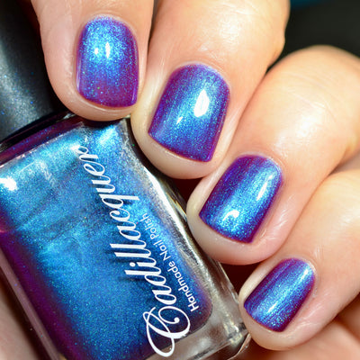 Multichrome shimmer Cadillacquer nail polish in Keep Doubting, swatched on five short squoval nails held alongside the bottle, shifting from deep mulberry-violet at rest to vivid cerulean blue under direct flash, with teal-green micro-sparkle visible, light skin tone, direct flash lighting, swatched by @caitlinswatches