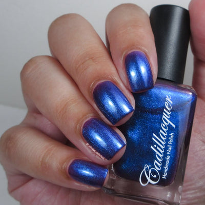 Shimmer multichrome Cadillacquer nail polish in Keep Doubting, swatched on eight short natural nails held alongside the bottle, showing a deep indigo-violet base packed with cobalt blue shimmer that shifts toward purple at lower angles, light skin tone, indoor warm lighting, swatched by @_always_polished_