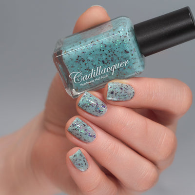 Cadillacquer Pastel Thoughts pastel teal purple chameleon iridescent crelly nail polish, bottle held with four-nail swatch, light skin, studio lighting