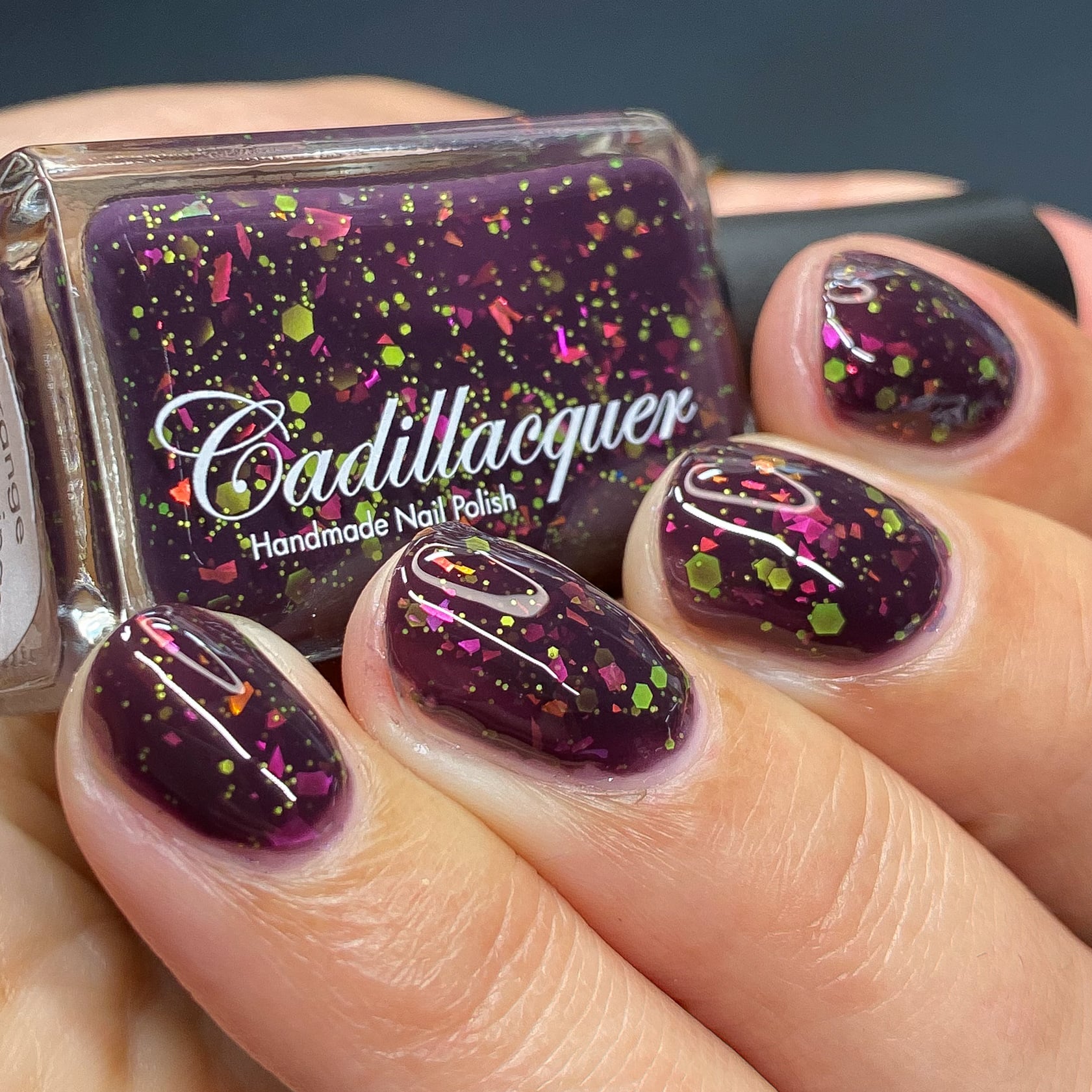 Thermal glitter and flake Cadillacquer nail polish in Strange Happenings, shown in a bottle-in-hand swatch with five short natural nails on light skin, the blackened eggplant purple base is loaded with neon yellow-green matte glitter in large hex and small dot sizes plus hot pink and orange iridescent flakes, photographed under studio lighting, swatched by @heeymercedes