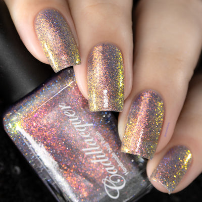 Holographic multichrome flake Cadillacquer nail polish in You Wished For This, shown on five squoval nails with a matching bottle, micro flakes shift from pinkish red-orange through copper to yellow-gold over a clear base with scattered blue and charcoal holographic glitter, light skin tone, direct flash lighting, swatched by @serpentine13