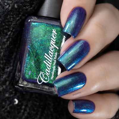 Multichrome holographic shimmer Cadillacquer nail polish in What Could Possibly Go Wrong?, bottle held in hand alongside four swatched nails showing a deep indigo base shifting through electric blue, teal-cyan, and vivid green depending on angle, bottle face dominated by green holographic shimmer with deep indigo visible at glass rim, light skin tone, studio lighting, swatched by @serpentine13