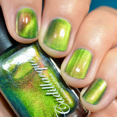 Holographic magnetic multichrome Cadillacquer nail polish in I Found A Place, swatched on five short natural nails showing a bright lime-green base with a warm copper-amber magnetic band and red-to-orange aurora shimmer flash, held alongside the polish bottle, light skin tone, direct flash lighting, swatched by @caitlinswatches