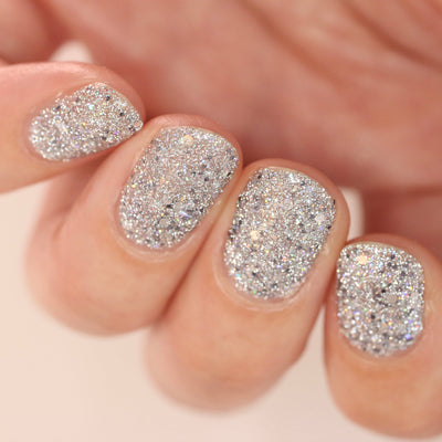 I Started the Dam Thing by Dam Nail Polish, densely packed silver glitter with multichrome holographic flashes, normal and flash studio views, on a light skin tone hand