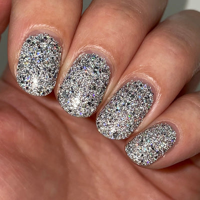 I Started the Dam Thing by Dam Nail Polish, full-coverage silver glitter with scattered rainbow holographic particles under direct flash, on a light-medium skin tone hand