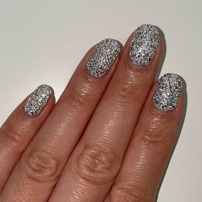 I Started the Dam Thing by Dam Nail Polish, dense silver reflective glitter with holographic micro-flakes in warm indoor light, on a light-medium skin tone hand