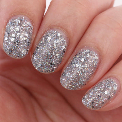 I Started the Dam Thing by Dam Nail Polish, silver and white mixed-size glitter over a sheer greige jelly base, normal and flash studio views, on a light skin tone hand