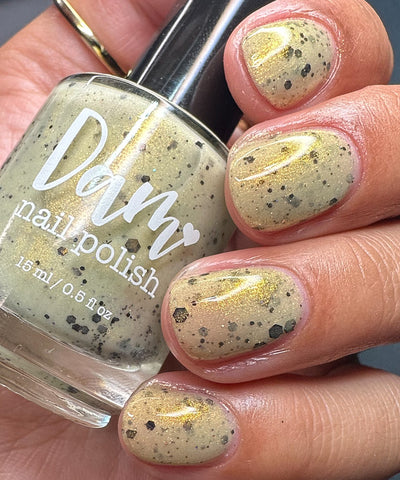 OG Devo-tea by Dam Nail Polish, warm tan crelly with gold shimmer and scattered black hex and dot glitters in daylight, bottle held in a light-medium skin tone hand