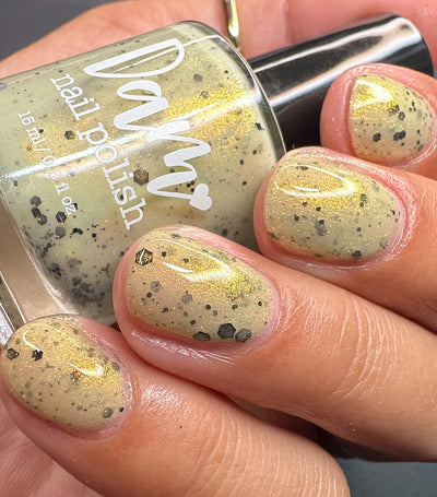 OG Devo-tea by Dam Nail Polish, warm tan crelly with gold shimmer and black hex plus fine particle glitters in bright daylight, bottle held in a light skin tone hand