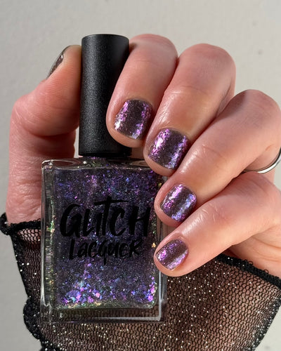 AVEN by Glitch Lacquer, soft black base with iridescent purple flakes and holo micro-glitter, bottle in hand on light skin tone