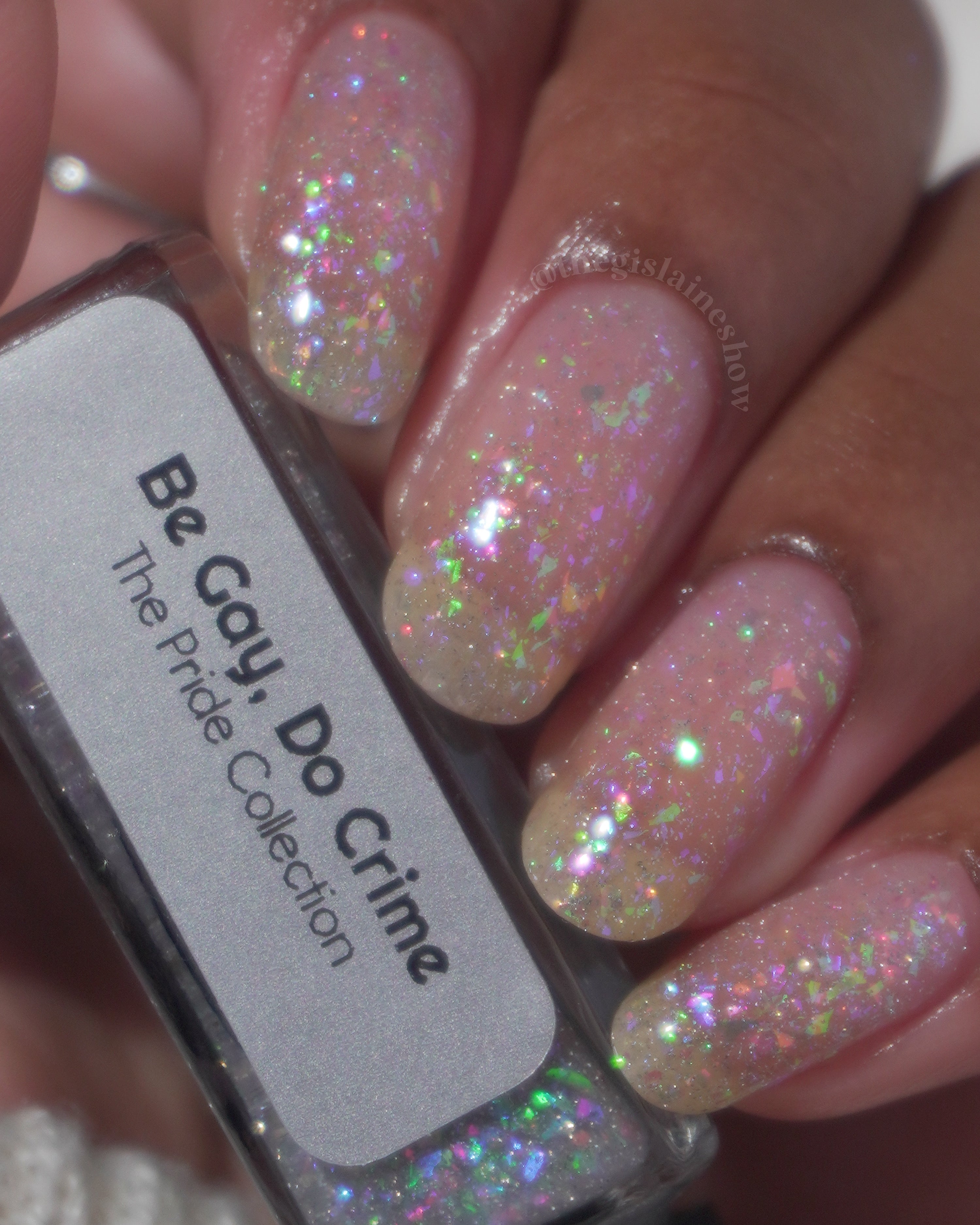 Be Gay, Do Crime by Glitch Lacquer, rainbow flake and holo glitter topper over sheer base, bottle in hand on medium-deep skin